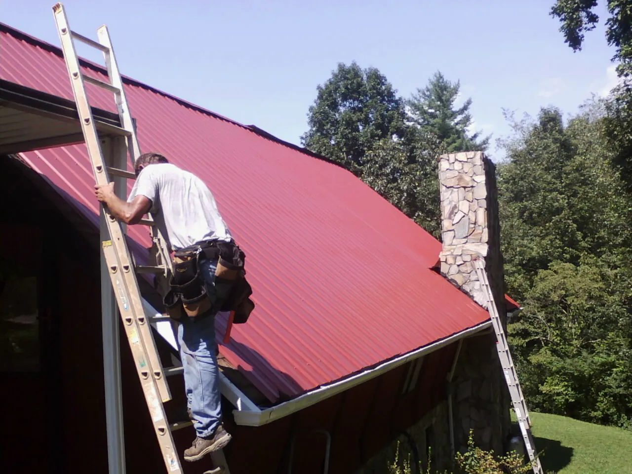 Roofer on a ladder beside a red metal roof and stone chimney — Soffit & Fascia Repair by Roofing Brothers in Grosse Pointe